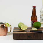 Timber & Tradition branded cutting board with cut limes, ginger beer, and Mezcal to prepare a Mezcal mule.