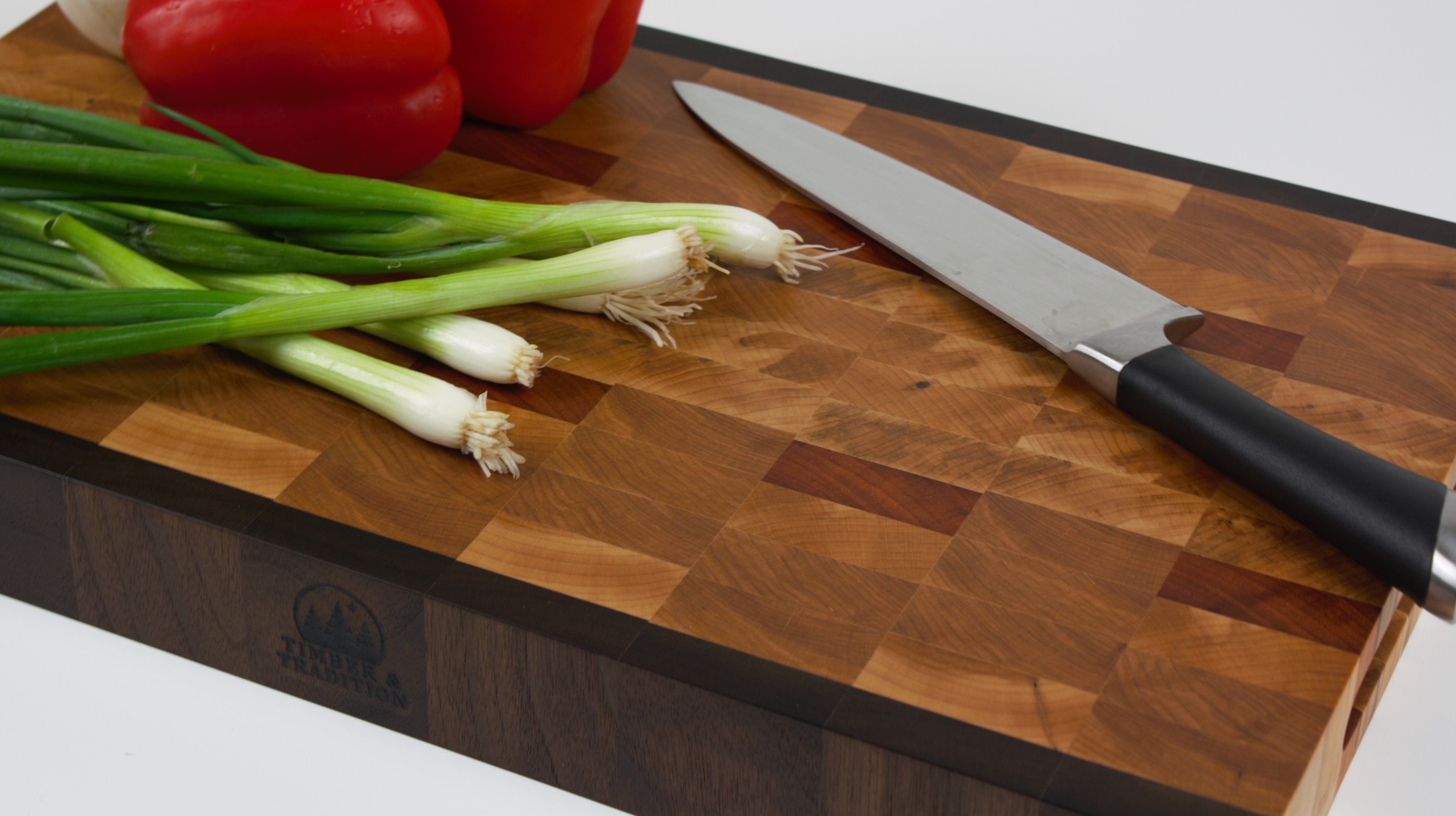 End grain cutting board made of rustic Hard Maple and accented with Tigerwood and Black Walnut. Red peppers and scallions lay on the cutting surface next to a chef's knife.